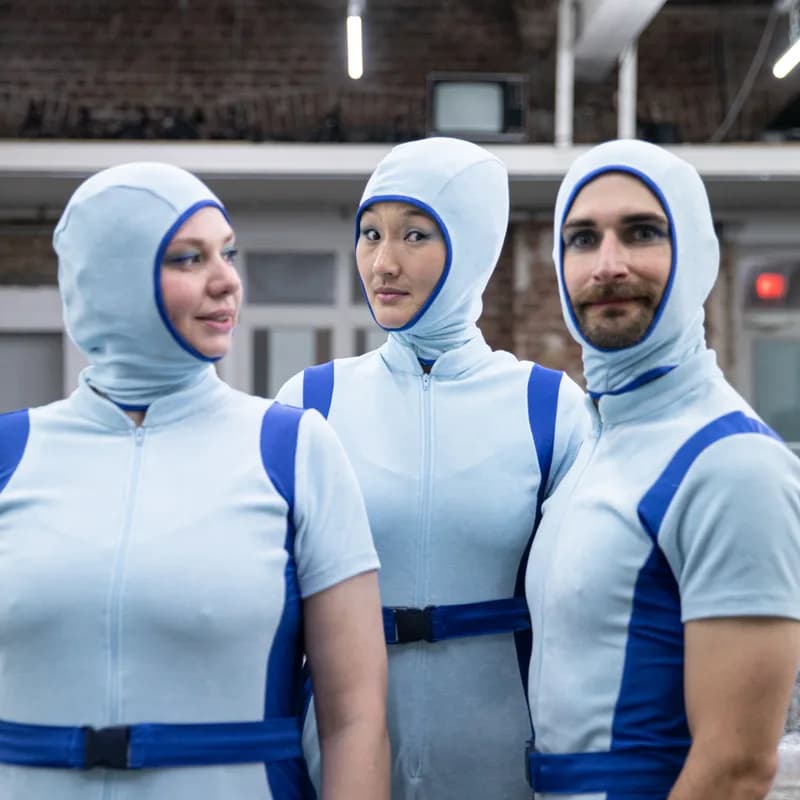 3 Personen in blauen kurzen Onesies mit aufgesetzten Kapuzen schauen in die Kamera.