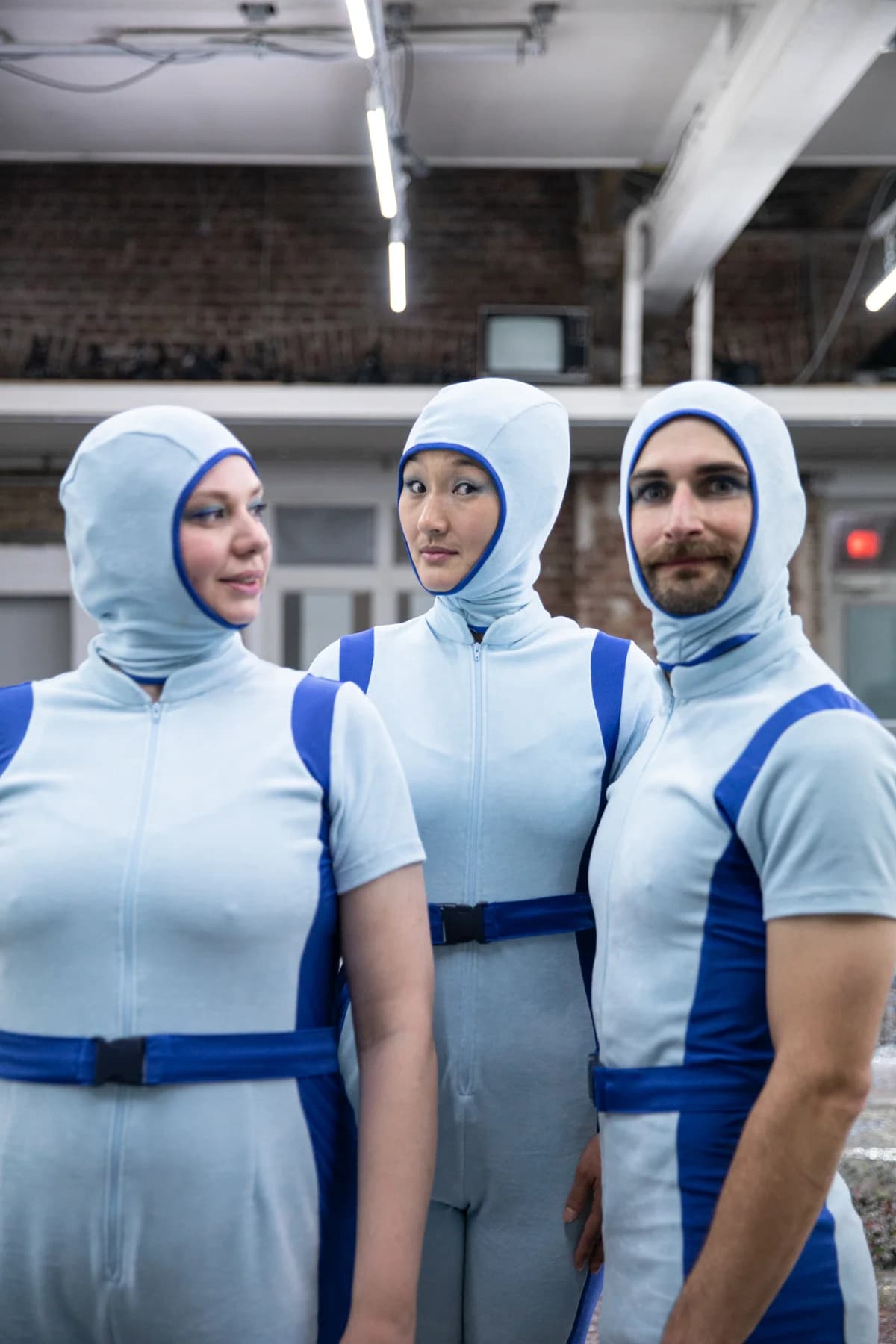 3 Personen in blauen kurzen Onesies mit aufgesetzten Kapuzen schauen in die Kamera.