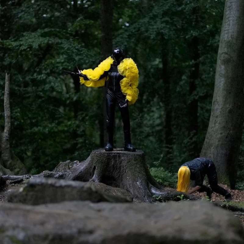 Zwei schwarz gekleidete Personen mit gelben Accessoires bewegen sich im Wald.