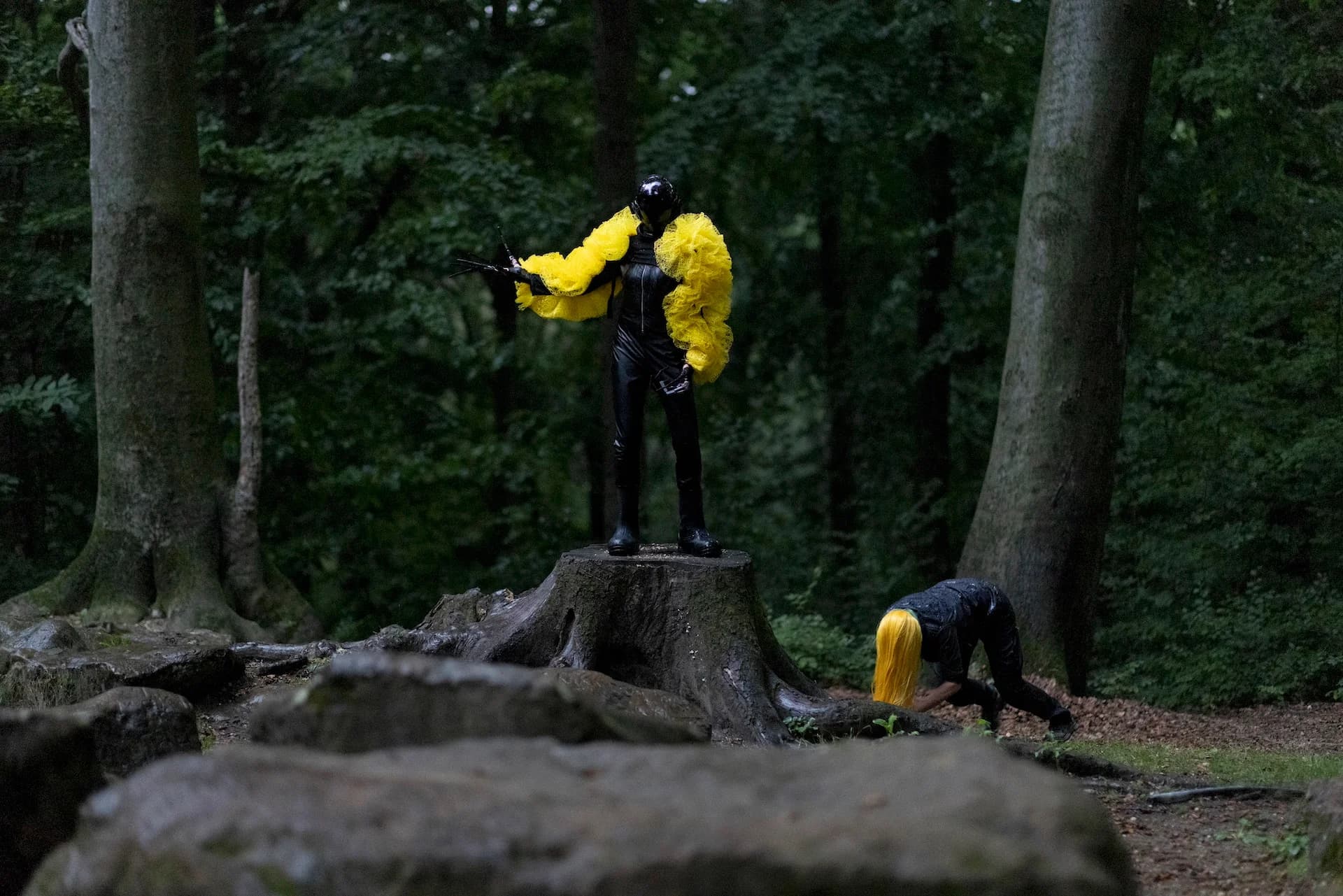 Zwei schwarz gekleidete Personen mit gelben Accessoires bewegen sich im Wald.