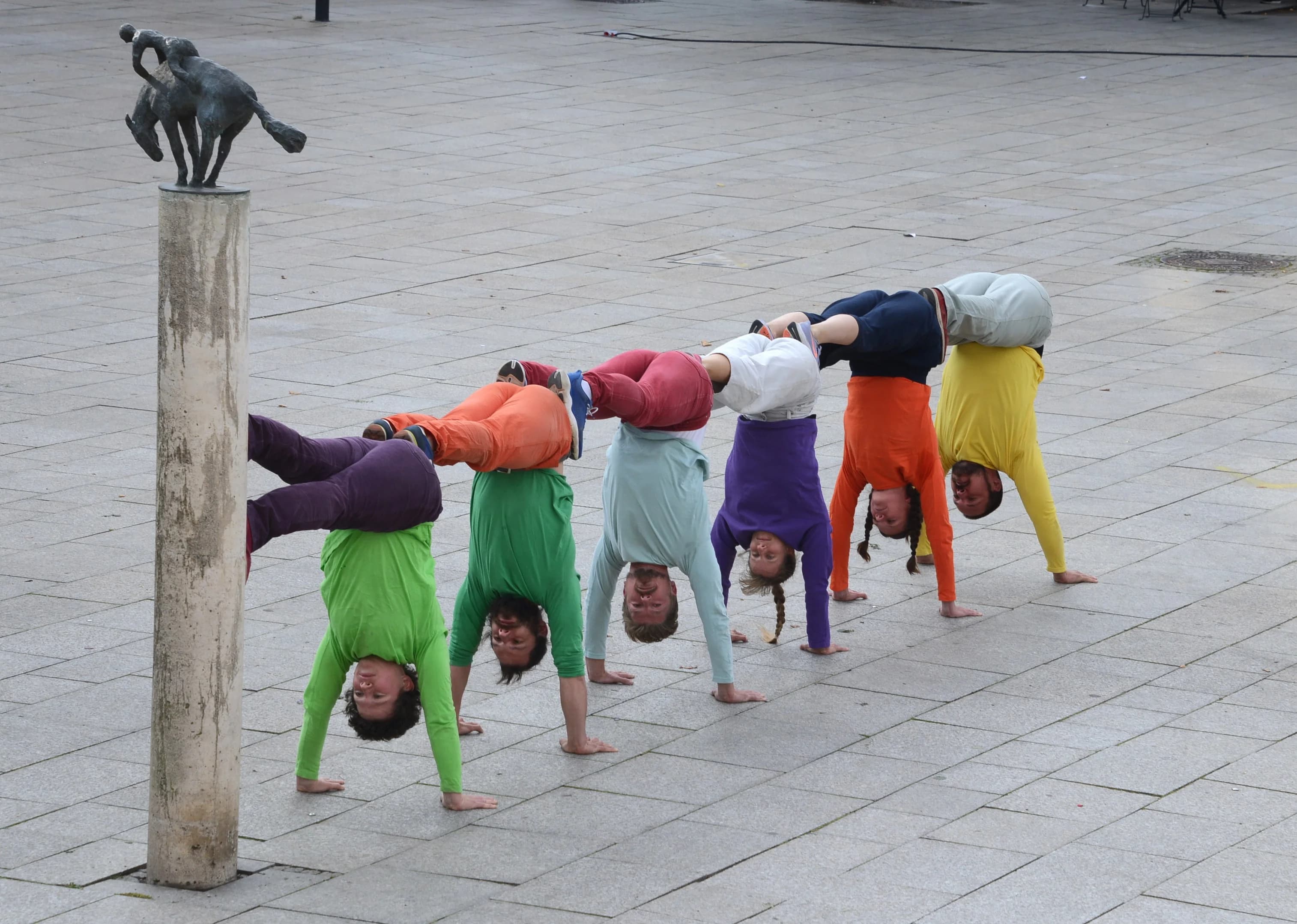 6 bunt gekleidete Personen im Handstand hintereinander.